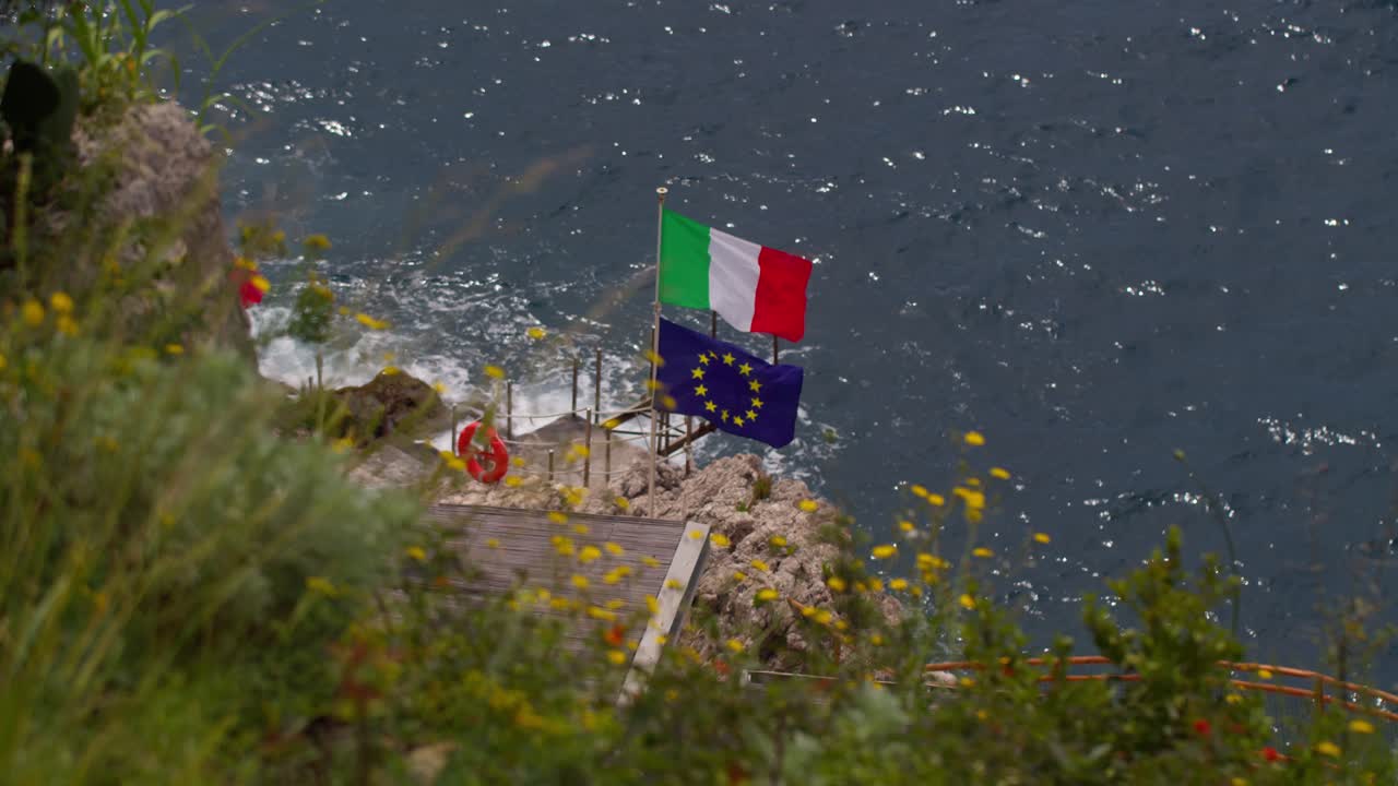 Italian and EU flags blowing in the wind with nature blurred in foreground in Taormina, Sicily, Italy (Italia)