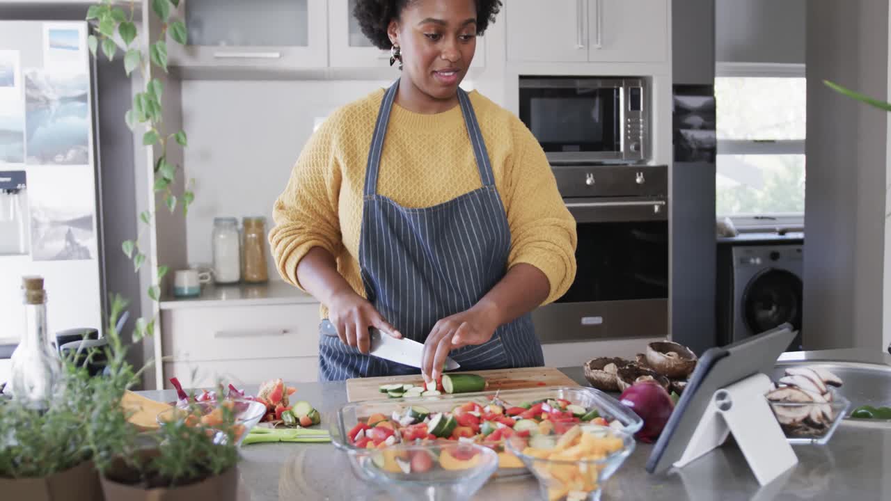 Happy african american woman preparing meal using tablet chopping vegetables in kitchen, slow motion
