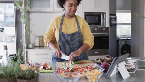 happy african american woman preparing meal using tablet chopping vegetables in kitchen, slow motion