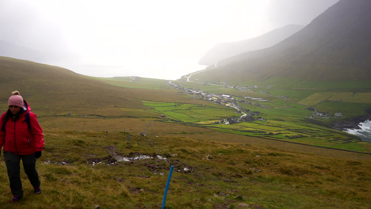 Donna che fa escursioni su una montagna nebbiosa, lasciandosi alle spalle il villaggio di Vidareidi, Isole Fær Øer