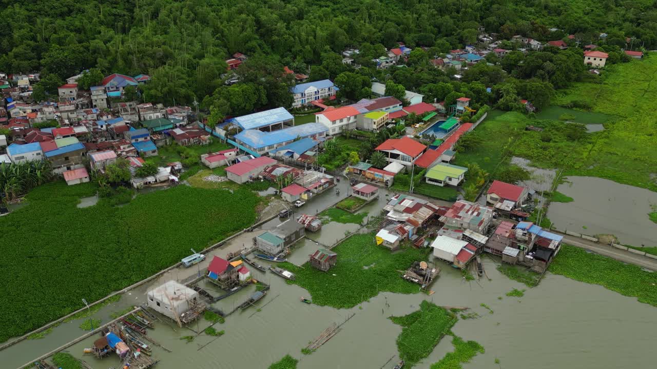 Wide aerial pass captures a lakeside Philippine town with submerged streets, vibrant rooftops, and lush greenery blending into overcast tropical skies