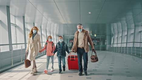 happy family wearing protective masks walking down the airport terminal corridor, carrying luggage, tracking shot