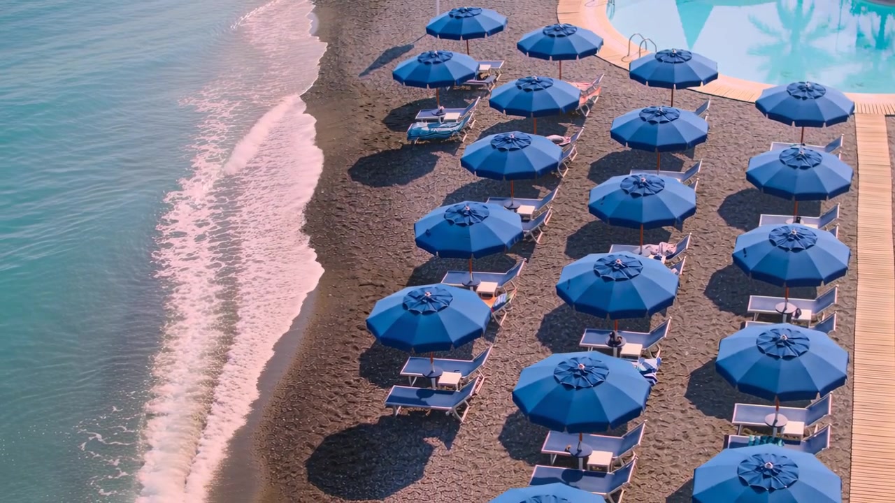 Drone che filma una spiaggia di un resort con file di ombrelloni blu e lettini con bagnanti, con le onde che si muovono