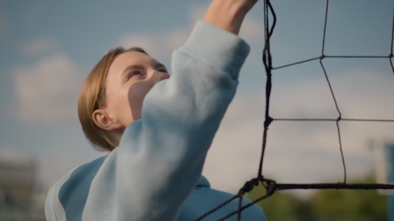 푸른 색의 후디를 입은 여성이 야외에서 배구망을 흔들고, 은 하늘 아래 스포츠 경기장에서 활동하는 배구 선수들의 흐릿한 배경으로