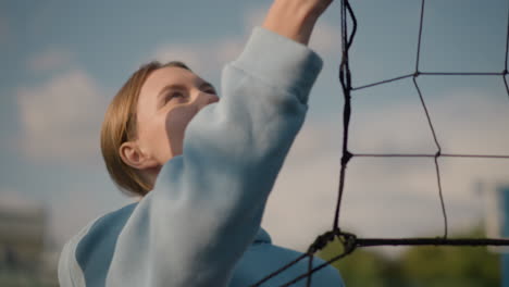 dama con capucha cian sacudiendo la red de voleibol al aire libre, con el fondo borroso de los jugadores de volleyball en acción en el campo deportivo bajo un cielo despejado