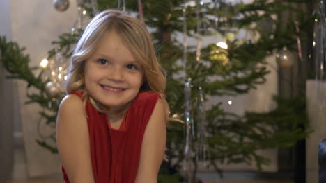 little blonde happy girl smiles with christmas tree behind her