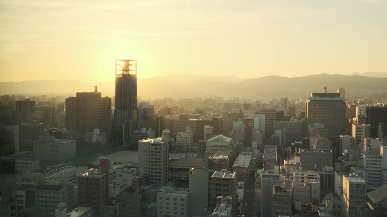 High-angle view of Hiroshima's skyline during golden hour. The city glows under warm sunset light, with distant mountains and modern high-rise buildings. Filmed from hotel window.