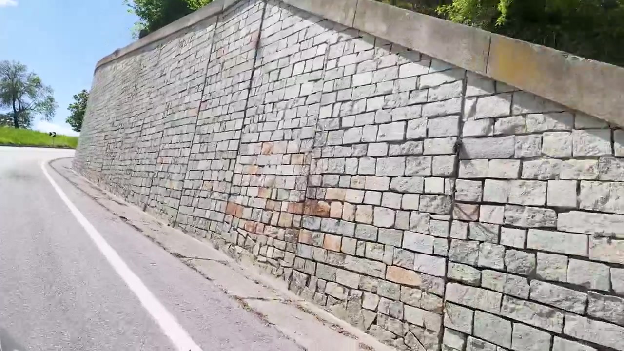Una telecamera panoramica lungo una strada asfaltata curva, oltre colline verdi e muri in pietra.