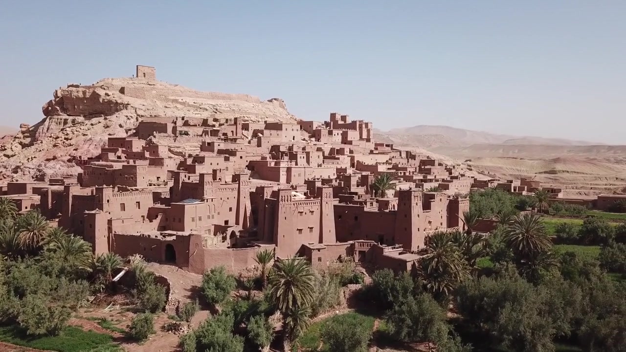 Ampia veduta della kasbah di Ait Benhaddou con un letto di fiume poco profondo e una persona solitaria che attraversa il primo piano. Iconico sito UNESCO a Ouarzazate che fonde la scala umana con il paesaggio desertico.