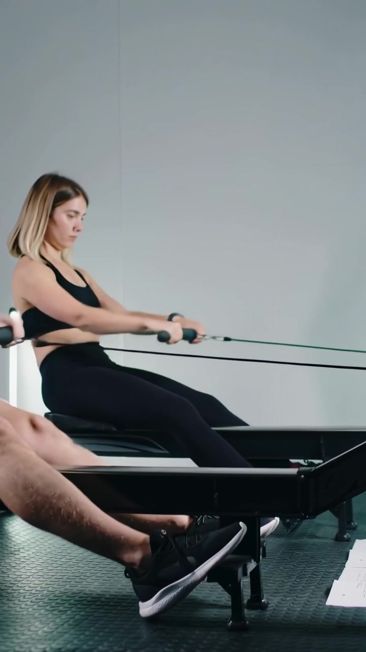 Vertical video: Starting couple rowing in sync on gym rowers with plan in athletic wear for fitness