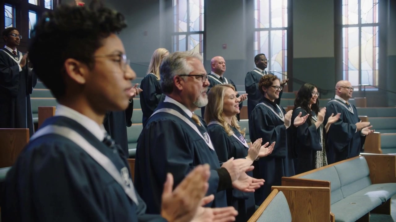 Coro in tuniche blu scuro con stole che si alza tra i banchi della chiesa, l'applauso scatenato dalla fine dell'assolo.