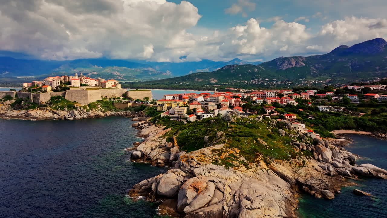 Aerial drone shot over the historic old town of Calvi, Corsica, France. Picturesque view of coastal town, landscape, fluffy clouds, and mountains in the background
