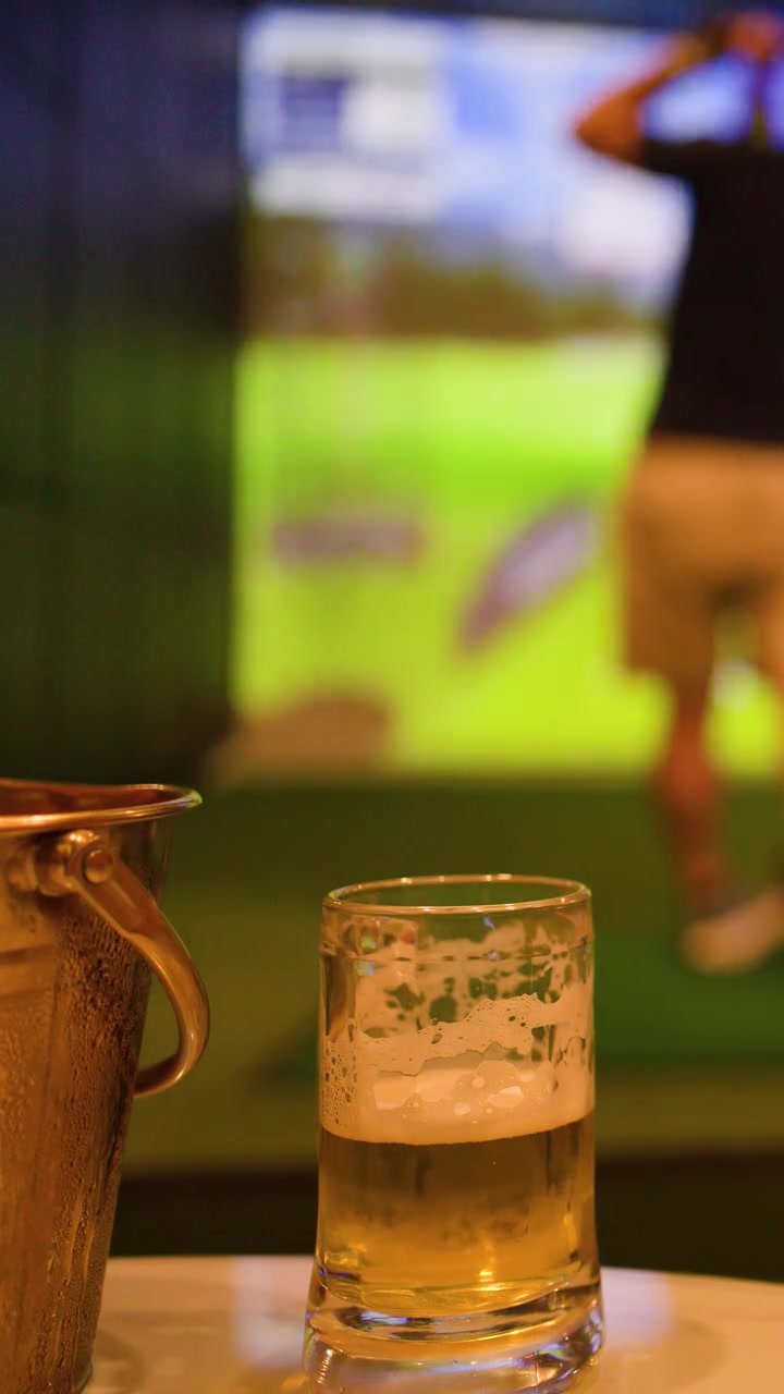 Un uomo esegue uno swing con una mazza da golf verso uno schermo simulatore dietro un bicchiere di birra fredda.