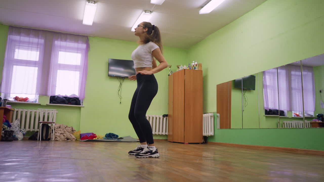 Young girl in white top and black leggings dancing in bright room with wooden floor and mirror wall, showing expressive body movement and energetic workout routine during practice session indoors