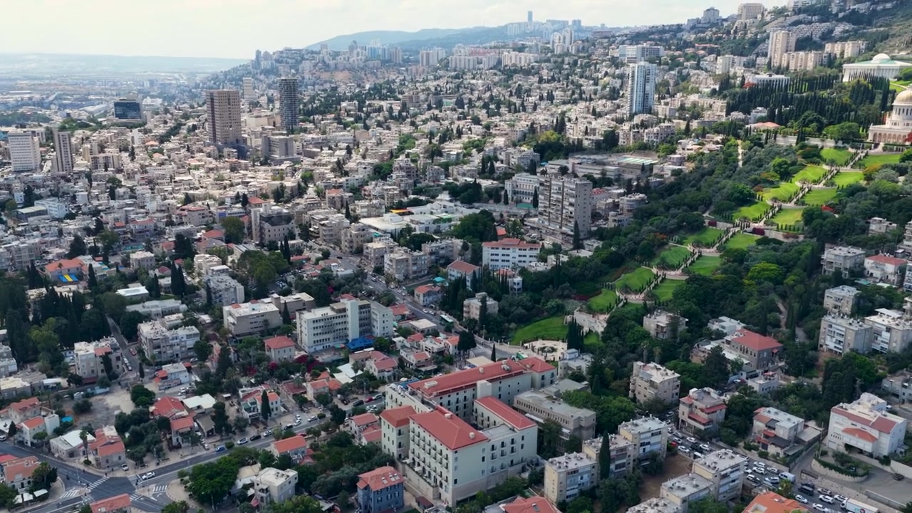 Riprese cinematografiche con drone che mostrano i giardini terrazzati Baháʼí sul Monte Carmelo, affacciati sulla densa espansione urbana e sui quartieri costieri della città di Haifa in una limpida giornata estiva.