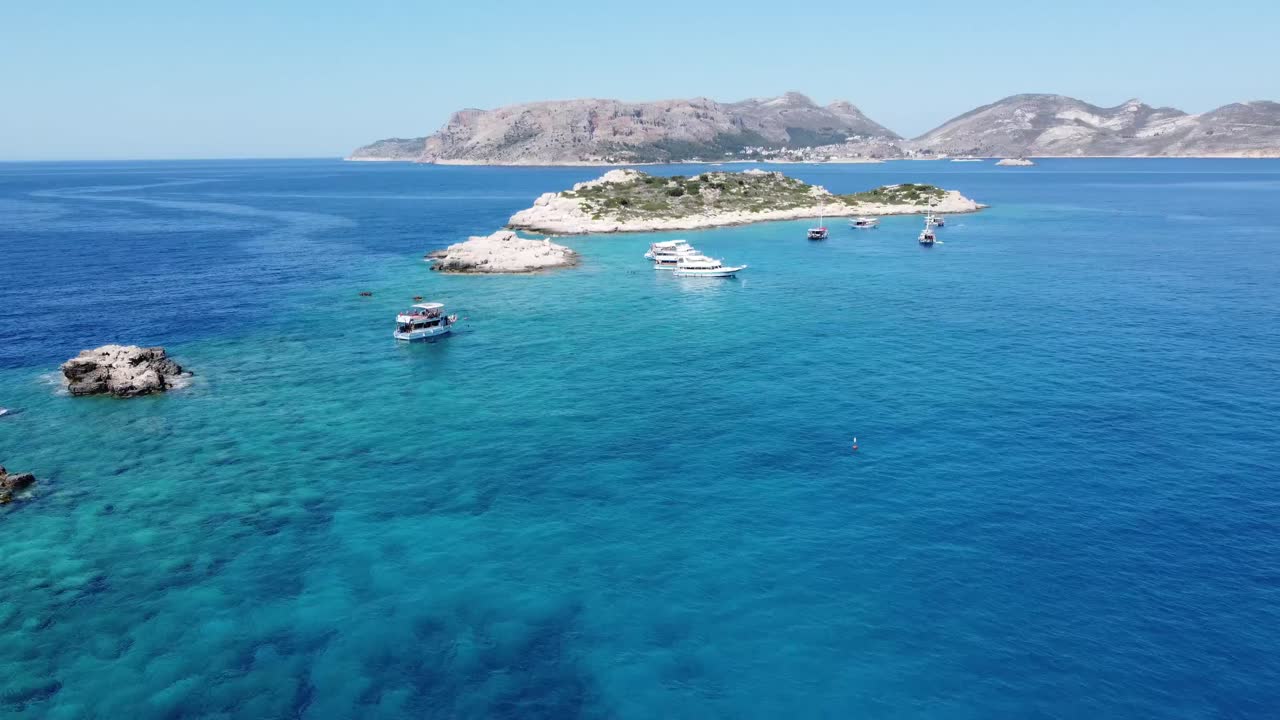 Drone video capturing boats gliding near rocky islets and a hilly coastline under clear skies off Kekova Island, Antalya, Turkey. The serene turquoise waters create a breathtaking landscape