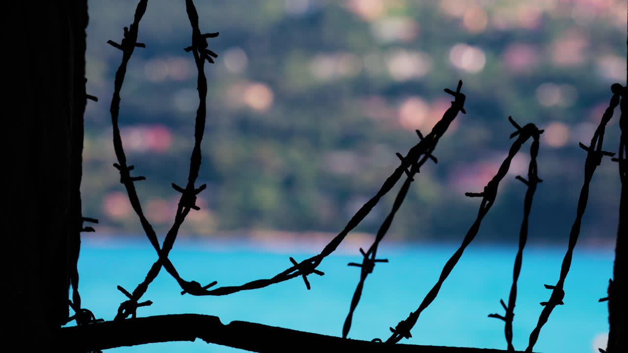 Close up of barbed wire loops along a small window with a blurred view of the sea and a town