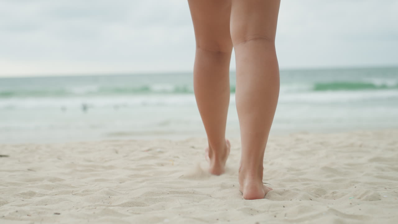 Gambe femminili a piedi nudi che camminano verso l'oceano sulla riva dell'isola, scena costiera contemplativa con onde leggere e orizzonte, luce cinematografica nuvolosa, umore riflessivo del viandante della spiaggia.
