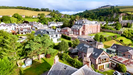 Drone camera glides above Rosemarkie, capturing residential homes, historic buildings, and lush greenery under bright daylight with smooth, steady movement