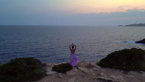 woman meditating in yoga position on a cliff overlooking the sea at sunset in ibiza. lovely aerial view flight fly reverse drone