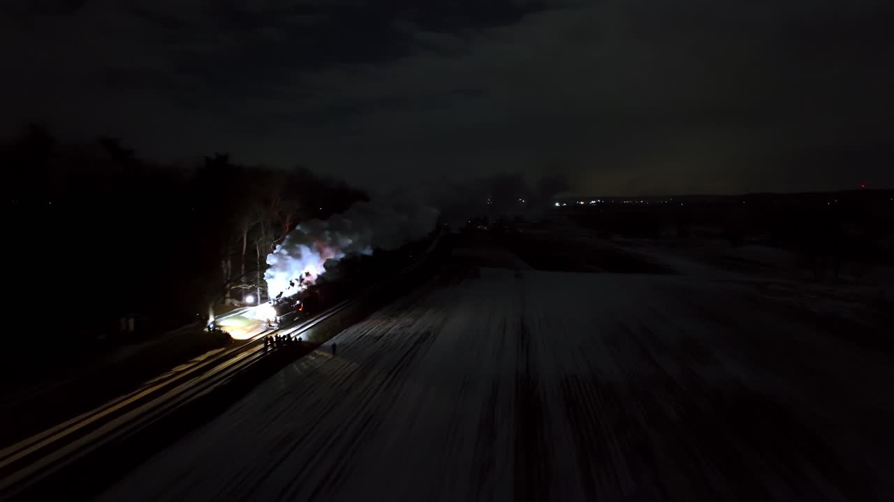 A train travels along dimly lit tracks in a snowy area at night. Smoke rises from the train as it moves, creating a dramatic contrast against the dark sky and winter scene.