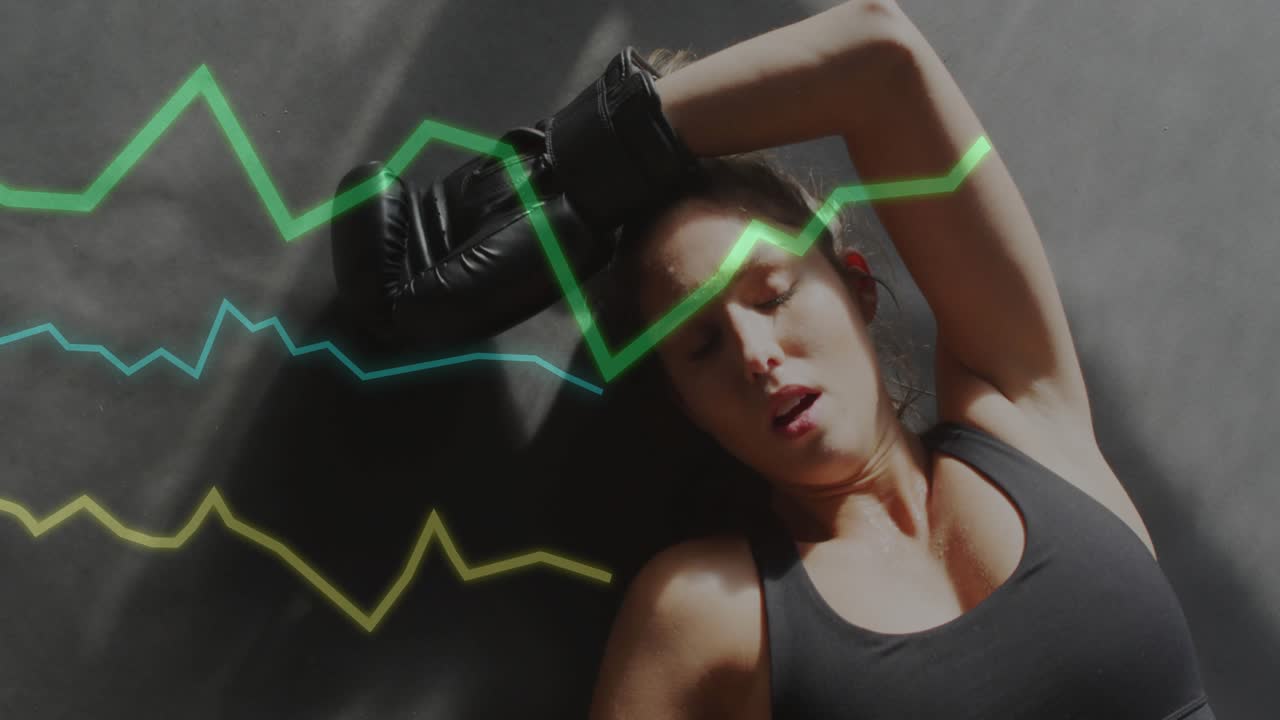 Fitness woman lying on mat responding to pulsing graph lines and raising gloved arm shielding face