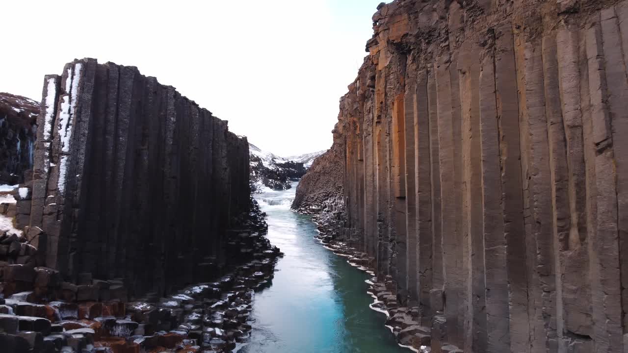 A breathtaking panoramic view showcases the unique natural beauty of Studlagil Canyon, where a serene, turquoise river flows between towering walls of geometrically perfect basalt columns