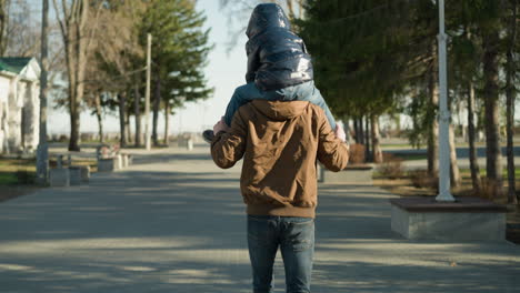 a father runs along a tree-lined paved path while carrying his child on his shoulders, the father holds the child's legs to maintain balance, both dressed in jackets