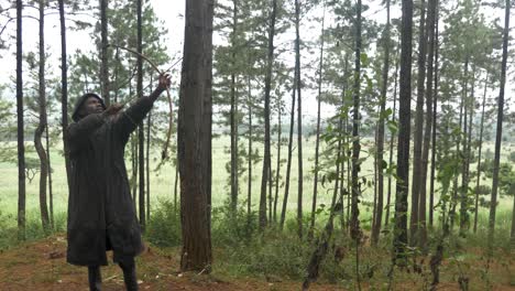 a hooded and cloaked african hunter in a pine forest shoots from his traditional wooden bow a arrow up into the trees