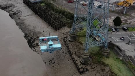 Historic-industrial-Newport-transporter-bridge-platform-moving-across-river-Usk-aerial-birdseye-high-orbit-left