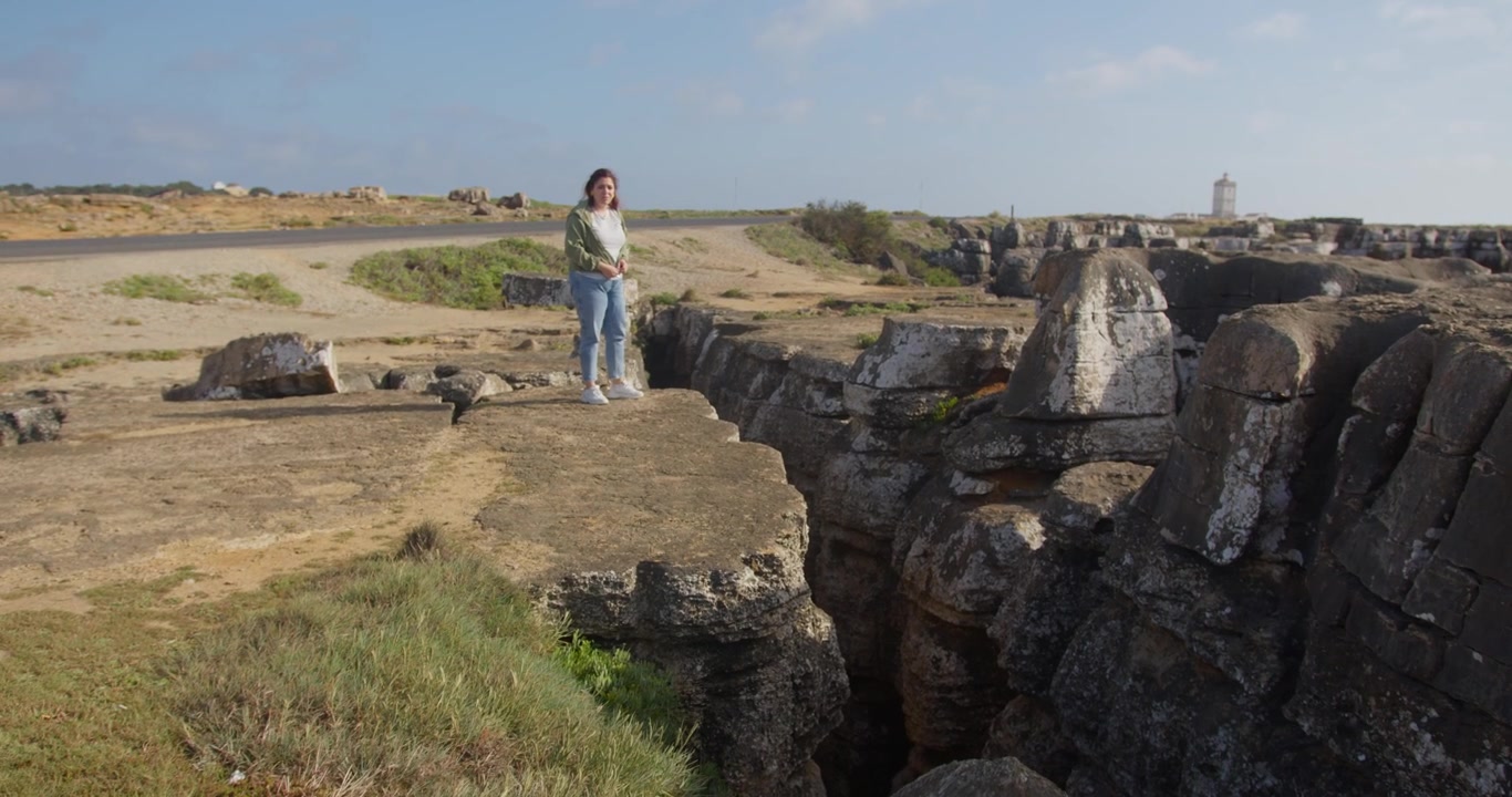 Ripresa aerea in 4K al rallentatore di una modella in piedi sul bordo di una singolare fessura nel terreno, che guarda giù dalla scogliera, circondata dal drammatico paesaggio di Peniche, Portogallo