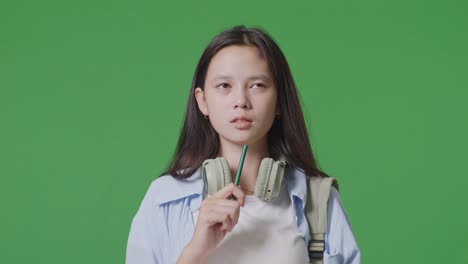 close up of asian teen girl student with a backpack taking note on notebook and thinking then raising her index finger while standing in the green screen background studio