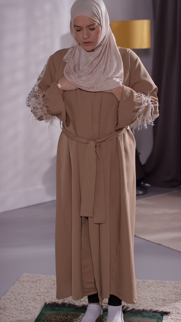 집에서 히자브를 입은 무슬림 여성이 기도 매트에 서서 기도하는 수직 비디오