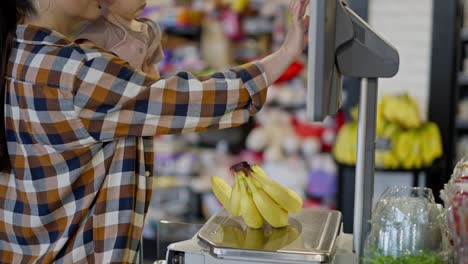 fotografia em close-up de uma mãe confiante com seu bebê nos braços pesando bananas em um relógio digital em um supermercado moderno