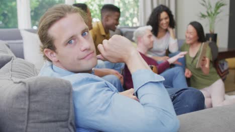 hombre caucásico sonriente sentado con un grupo diverso de amigos felices socializando en la sala de estar