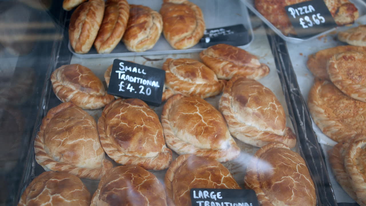 Cornish pasties displayed behind a bakery window include traditional and cheese topped varieties with hand written price labels on the metal trays