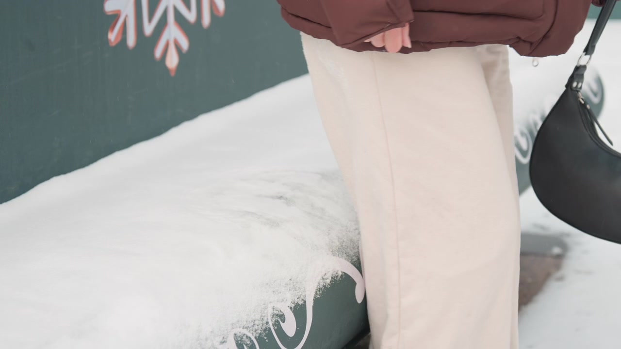Bambino o persona minuta si sposta dalla posizione seduta a quella eretta su una panchina coperta di neve, mentre indossa una giacca a piumino trapuntata marrone e pantaloni beige chiaro, la mano afferra la tracolla di una borsa scura mentre il corpo si gira e il peso si sposta.