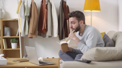 bearded young man sitting on couch and talking on video call on laptop while eating noodles from takeout box