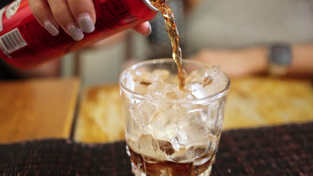 A hand pours cola from a red can into a clear glass filled with ice cubes on a wooden table, under soft indoor lighting with shallow depth of field