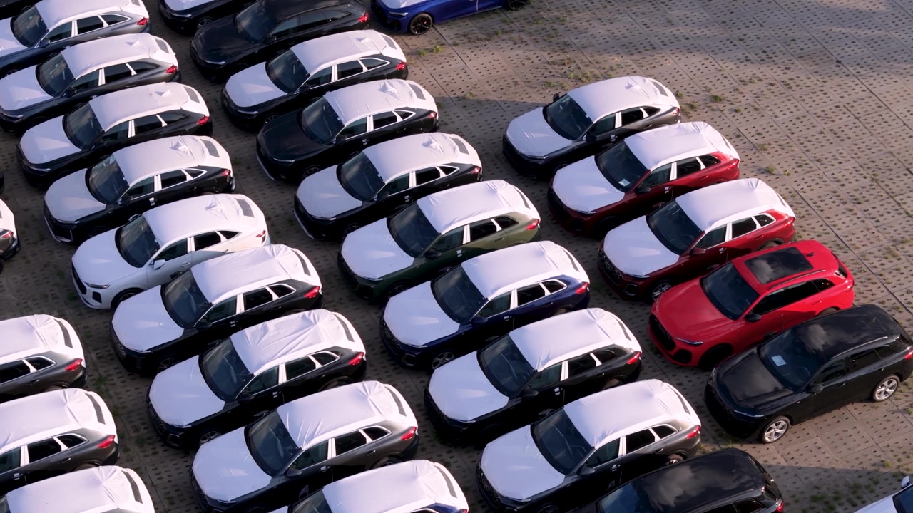 Ripresa drone dall'alto che mostra file di auto nuove parcheggiate presso una struttura industriale di logistica e distribuzione, evidenziando la catena di approvvigionamento automobilistica