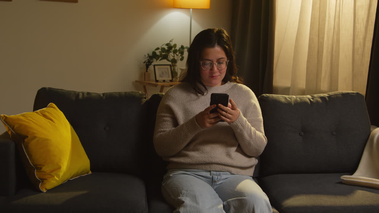 mujer joven pasando la noche en casa sentada en el sofá con el teléfono móvil desplazándose por internet o redes sociales 3