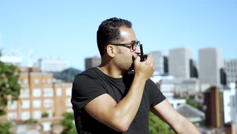 a man in a black t-shirt uses a walkie-talkie on a rooftop with a clear view of an urban skyline featuring buildings and greenery, showcasing communication in a city setting