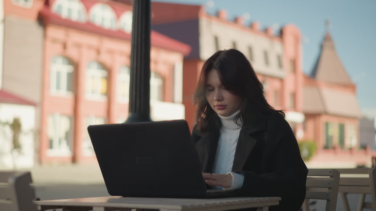 쇼핑몰에서 야외에 앉아 노트북에서 집중적으로 일하는 프리랜서, 배경에는 흐릿한 도시 건물, 편안한 작업 공간 환경에는 세련된 의상, 나무 테이블, 자연 빛이 있습니다.