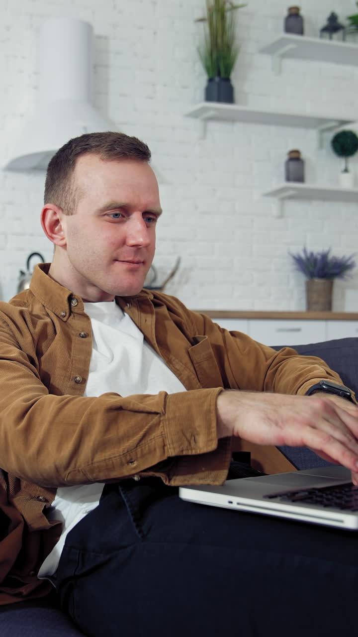 Young man working at home. Relaxed man laying on a sofa and typing on a laptop. Student studying online at home during quarantine. Remote job. Vertical video