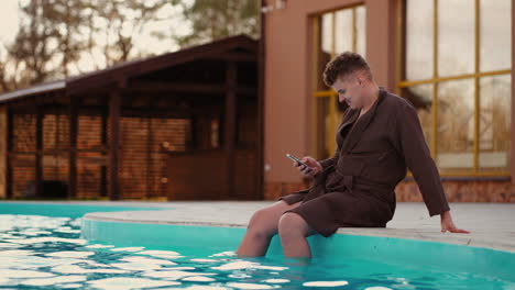 relaxed man is resting in outdoor park swimming pool surfing social media by modern smartphone