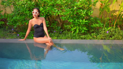 Chic-young-asian-woman-in-swimsuit-sitting-on-swimming-pool-border,-enjoying-hot-weather-on-her-tropical-vacation,-full-frame