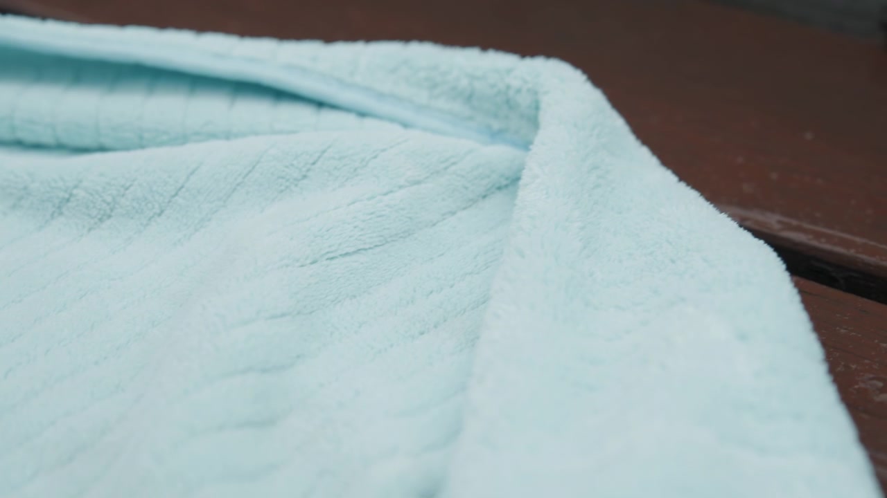 Close up of light blue towel with visible texture draped over weathered wooden surface. Soft lighting highlights fabric details and subtle color variations creating calm visual interest