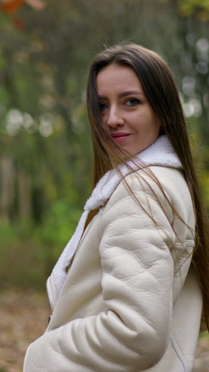 Long-haired brunette woman standing in the autumn park turns around to the camera. Lady looks into camera slowly approaching to her face. Blurred backdrop.