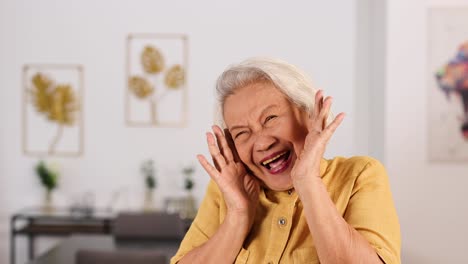 asian grandmother in yellow shirt expresses shyness and joy with playful hand gestures in a bright, modern living room with soft natural lighting