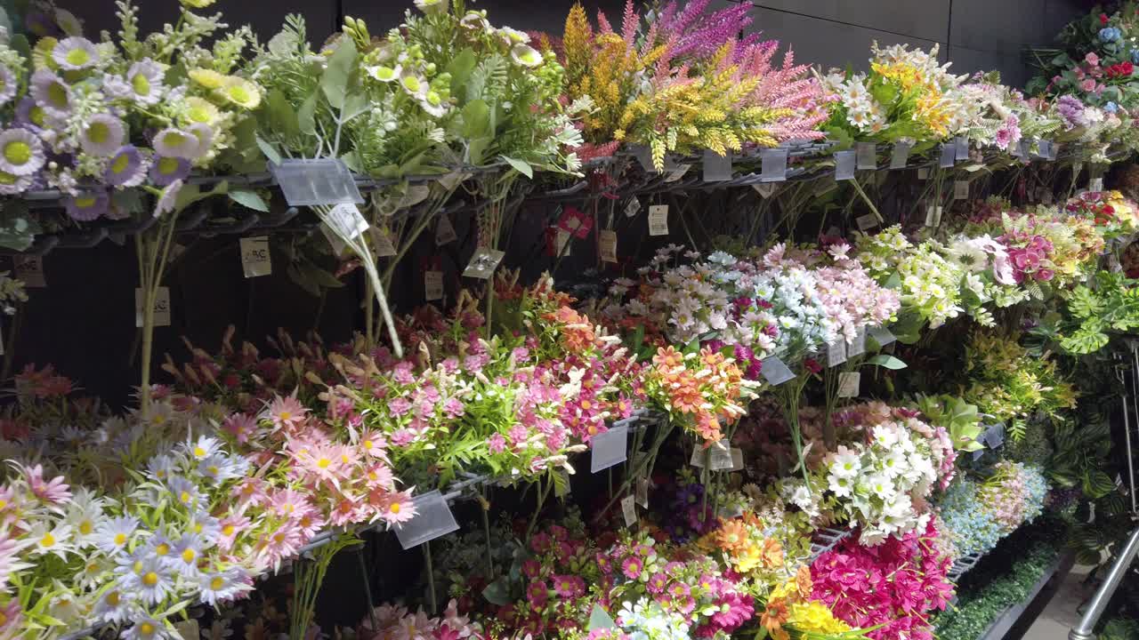 Inquadratura con carrellata a destra che mostra un fitto negozio di fiori con bouquet misti, fiori artificiali, colori vivaci e composizioni decorative sotto illuminazione interna